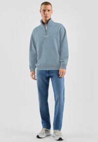Felpa azzurro chiaro con cerniera a quarto, polsini e orlo a costine, abbinata a jeans blu e sneakers grigie. Design minimalista con logo.