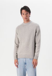 Hellgrauer gestrickter Pullover mit rundem Halsausschnitt, gerippten Ärmelbündchen und Saum, versehen mit einem dezenten Markenzeichen auf der Brust. Kombiniert mit hellblauen Jeans.