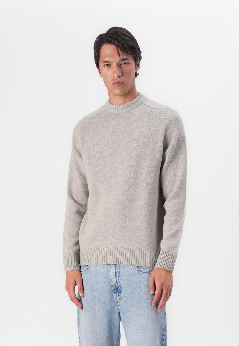 Hellgrauer gestrickter Pullover mit rundem Halsausschnitt, gerippten Ärmelbündchen und Saum, versehen mit einem dezenten Markenzeichen auf der Brust. Kombiniert mit hellblauen Jeans.