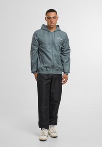 Šedá lehká bunda s kapucí, s předním zipem, postranními kapsami a elastickými manžetami, spárovaná s černými kalhotami a krémovými teniskami.