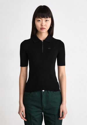 Polo shirt nero a coste con chiusura a mezza zip, maniche corte e un logo discreto. Indossato con pantaloni verdi.