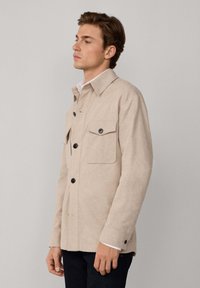Chaqueta beige hecha de una mezcla de lana, con un cuello puntiagudo, cierre frontal de botones, dos bolsillos en el pecho y puños abotonados.