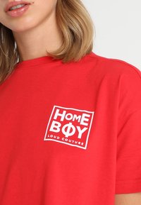 Camiseta de algodón roja con un logotipo gráfico blanco "HOME BOY LOUD COUTURE" en un diseño rectangular en el área del pecho izquierdo.