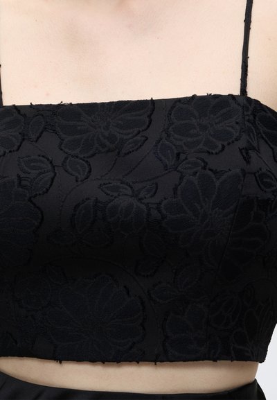 Haut court noir avec fines bretelles présentant des motifs floraux texturés porté sur peau nue.