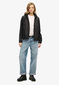 Veste légère noire, fermeture éclair à l'avant, capuche ajustable, associée à un jean large bleu clair et des bottes noires à lacets.