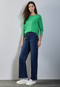 Vihreä pitkähihainen neule on yhdistetty tummansinisiin leveälahkeisiin farkkuihin ja valkoisiin lenkkikenkiin. Asu on rento ja neuleen pinta on sileä.