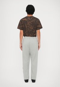 Camisa de manga corta marrón con patrón y textura abstracta, combinada con pantalones de chándal gris claro. Persona de espaldas, lleva zapatos negros.