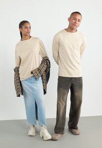 Beige long-sleeve shirts on two models, one with a plaid shirt, light blue denim skirt with a slit, and the other in dark jeans and clogs.