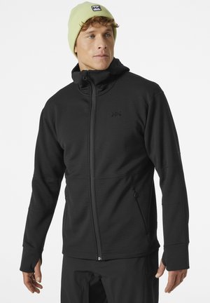 Man wearing black zip-up jacket with thumb holes and light green beanie, looking slightly right on plain white background.