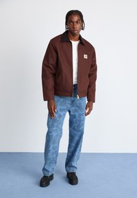 Brown zip-up jacket with black collar, white shirt underneath, blue denim pants featuring a subtle pattern, and black shoes.