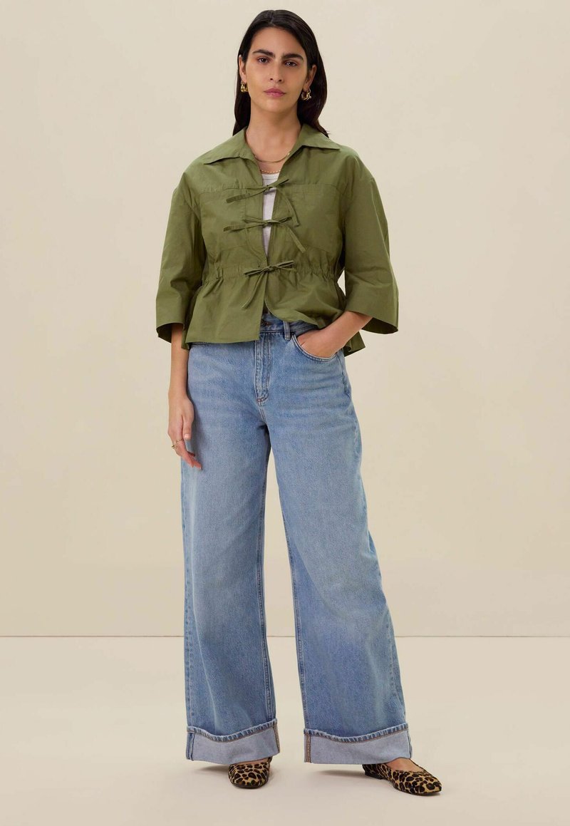 Veste cropped couleur olive avec des liens à l'avant, associée à un jean bleu à jambes larges et des ballerines à imprimé léopard. Texture de tissu lisse et coupe décontractée.