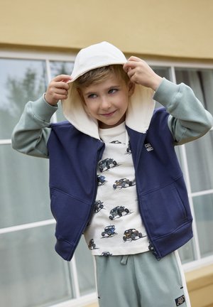 Gilet navy con zip e cappuccio chiaro, indossato sopra una camicia color crema con illustrazioni di automobili. Completano l'outfit pantaloni della tuta grigi.