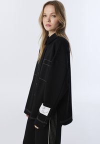 Czarna oversize koszula z białym przeszyciem i dwiema kieszeniami zapinanymi na guziki. Zawiera metkę na rękawie oraz klasyczny kołnierzyk.