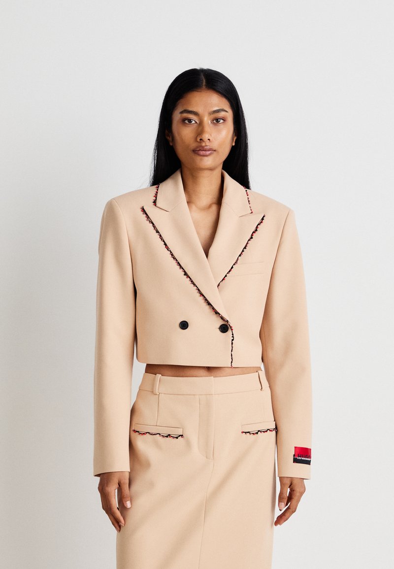 Beige Blazer mit schwarzen Knöpfen, mit kontrastierenden roten und schwarzen Akzenten entlang der Revers, kombiniert mit einem ähnlich gestylten beigen Rock mit Taschen.
