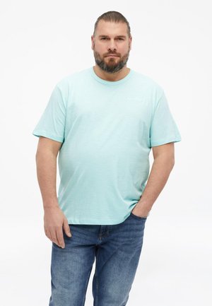 Mężczyzna z brodą i związanymi włosami, ubrany w jasnoniebieski t-shirt i dżinsy, stojący z jedną ręką w kieszeni na białym tle.
