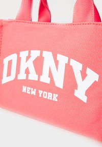 Roza tkaninska torba z dvema kratkima ročajema, ki prikazuje belo besedilo "DKNY" in "NEW YORK", z teksturirano površino.
