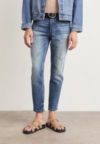 Persona vistiendo unos vaqueros azules desgastados con un ligero desgaste, una chaqueta de denim y sandalias beige, de pie sobre un suelo de hormigón.