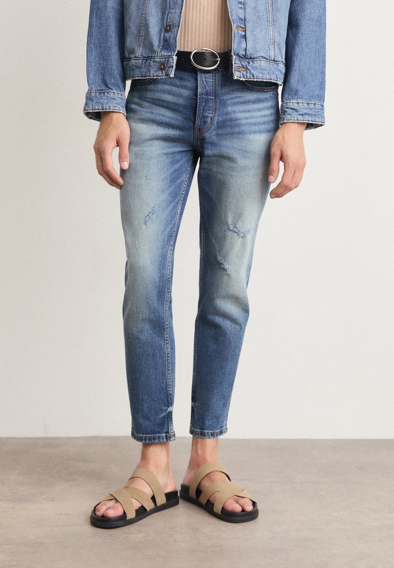 Persona vistiendo unos vaqueros azules desgastados con un ligero desgaste, una chaqueta de denim y sandalias beige, de pie sobre un suelo de hormigón.