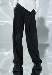 Isik, kes kannab musta triibulisi laia säärega pükse, käed taskutes, valget särk ja mustad paksud platvormkingad peegeldaval põrandal.