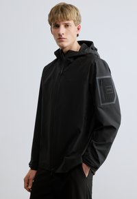 Fekete vízálló dzseki kapucnival, elülső cipzárral, oldalzsebekkel és könyökvédő részlettel. Simas textúra, minimalista design és logó hangsúly.