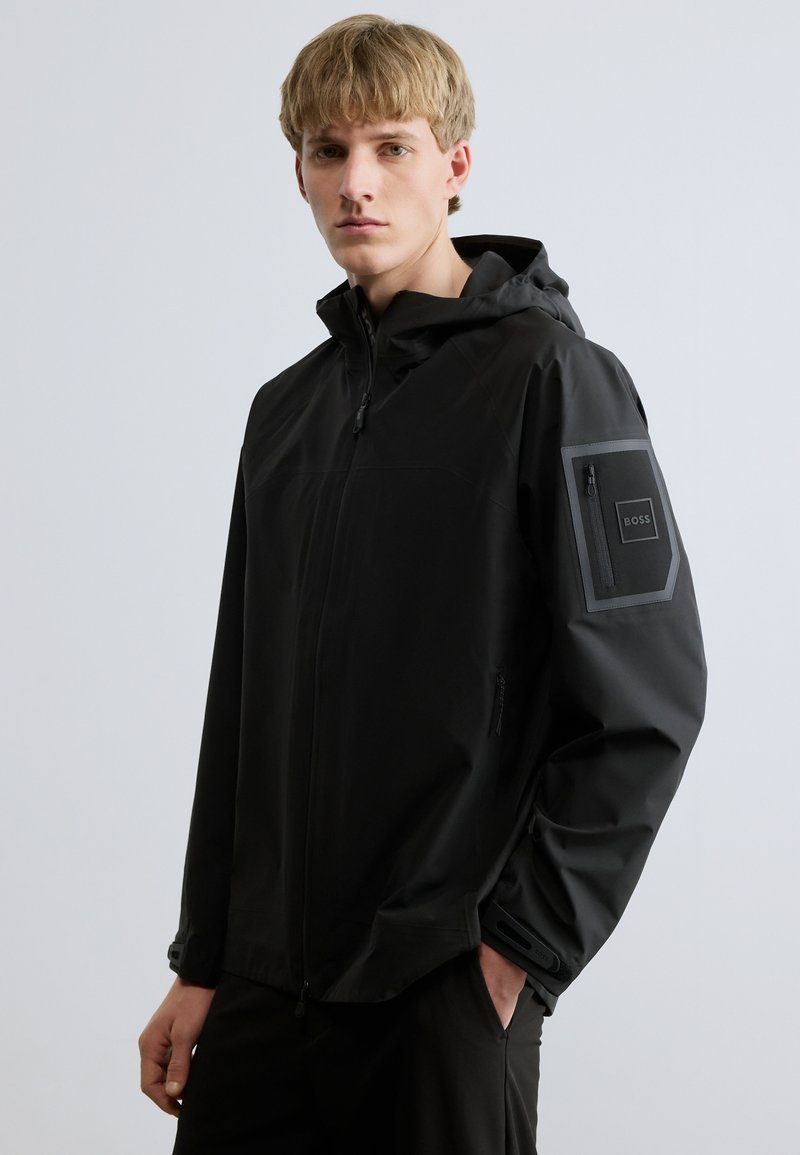 Fekete vízálló dzseki kapucnival, elülső cipzárral, oldalzsebekkel és könyökvédő részlettel. Simas textúra, minimalista design és logó hangsúly.