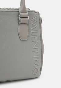 Bolso de cuero gris con superficie texturizada, logo en relieve y herrajes de plata. Cuenta con asas cortas y un cierre con cremallera.