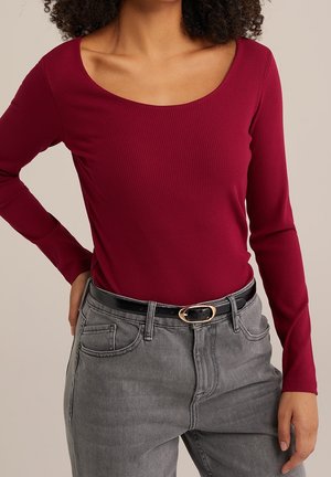 Vrouw draagt een geribbelde, bordeauxrode top met lange mouwen en een ronde hals, gecombineerd met een grijze jeans met hoge taille en een zwarte riem.