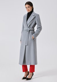Cappotto grigio lungo con cintura, colletto a revers e tasche. Indossato sopra un dolcevita nero e abbinato a pantaloni rossi e tacchi neri.