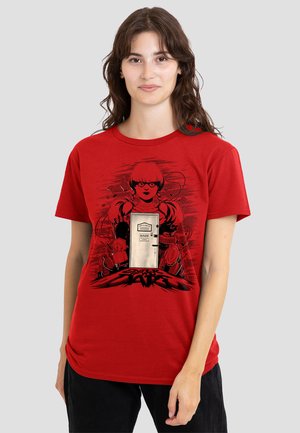 Young woman with dark wavy hair wears red T-shirt featuring a stylized doll with pigtails standing behind a door labeled "Danger".