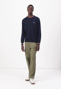 Navyblauwe gebreide trui met contrasterende oranje geribde halsrand, gecombineerd met olijfgroene broek en beige schoenen met bruine accenten.