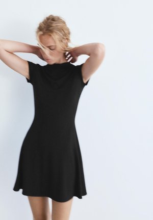 Žena s blond vlasmi si upravuje vlasy, oblečená v čiernych šatách s krátkymi rukávmi a dĺžkou po kolená, na jednoduchom bielom pozadí.