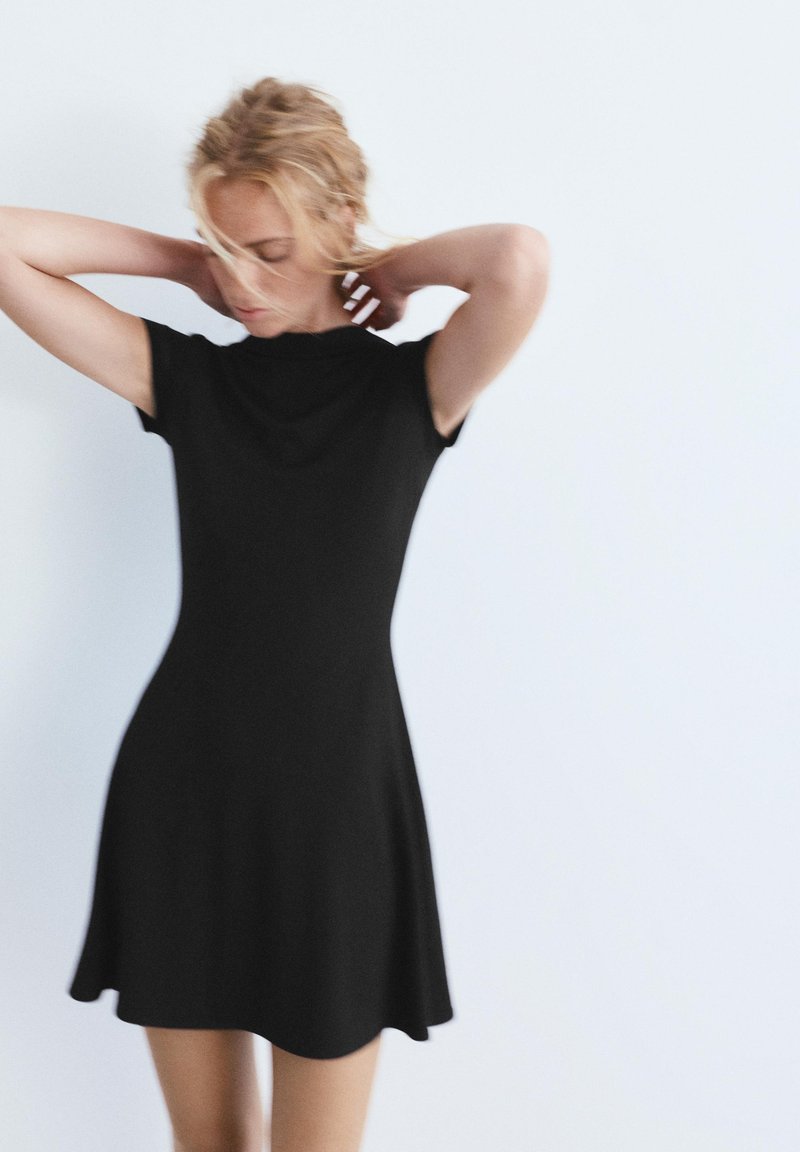 Szőke hajú nő, aki a haját igazítja, fekete rövid ujjú, térdig érő ruhát visel, egyszerű fehér háttér előtt.