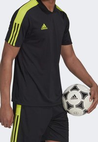 Svart idrottströja med gröna accenter och tre ränder på ärmarna, tillsammans med svarta shorts. Spelaren håller i en svartvit fotboll.