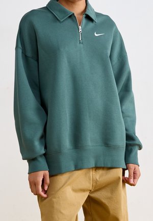 Personne portant un pull vert à manches longues avec une demi-fermeture éclair et un logo swoosh blanc, associé à un pantalon marron clair.
