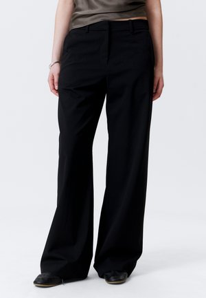 Person wearing black wide-leg pants, black shoes, and an olive green cropped top against a plain white background.