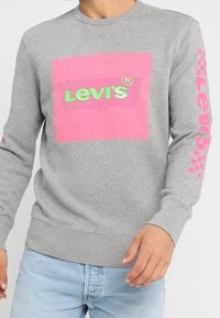 Sweatshirt gris avec un graphisme carré rose portant "Levi's" en vert, et un texte rose le long de la manche gauche. Tissu doux avec des poignets côtelés.