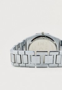 Ρολόι από ανοξείδωτο ατσάλι με μπρασελέ αποτελούμενο από ορθογώνιους συνδέσμους, οκταγωνική κάσα και στρογγυλό καντράν με χαραγμένες λεπτομέρειες της μάρκας στο πίσω μέρος.