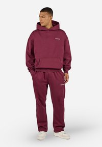 Man wearing burgundy hoodie and matching sweatpants, standing with hands in pockets, looking left, against white background.