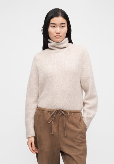 Bézs garbó pulóver puha kötött anyagból, hosszú ujjakkal és laza fazonnal, barna zsinóros nadrággal párosítva.