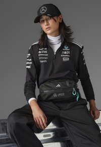 Schwarze Zip-Jacke mit weißen Akzenten und Logo-Drucken. Schwarzer Bauchtasche mit türkisfarbenem Reißverschluss, über schwarze Hosen getragen. Schwarze Kappe.