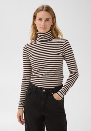 Femme aux cheveux blonds portant un col roulé rayé noir et blanc et un pantalon noir, debout avec une main dans la poche devant un fond uni.