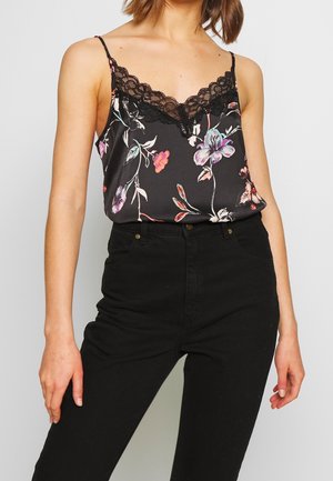 Kobieta ubrana w czarną satynową koszulkę na ramiączkach w kwiatowy wzór z koronkową lamówką i cienkimi ramiączkami, zestawioną z czarnymi spodniami z wysokim stanem.