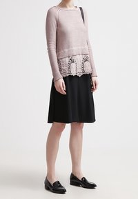 Maglione rosa chiaro con sovrapposizione in pizzo testurizzato sull'orlo, abbinato a una gonna nera a forma di A e mocassini neri. Materiali lisci, design aderente.