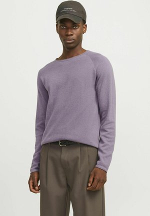 Suéter largo de manga larga texturizado en color púrpura con cuello redondo, combinado con pantalones claros de corte plisado en color marrón y una gorra negra con texto blanco.