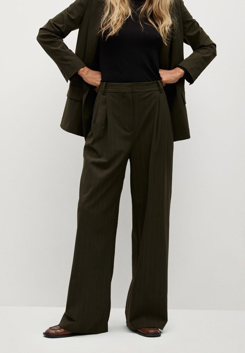 Femme portant un pantalon large à fines rayures vert olive, un blazer assorti, un top noir et des chaussures marron, debout les mains sur les hanches.