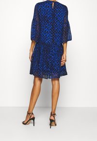 Blå leopardmönstrad klänning med transparent tyg, trekvarts ärmar med klockform och detalj med nyckelhål på ryggen, i kombination med svarta remmar på högklackade skor.