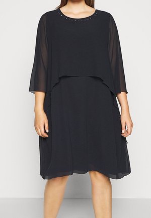 Robe noire à couches, longueur genou, avec manches trois-quarts transparentes et petits clous décoratifs autour du décolleté portée par une personne.