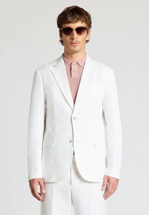 Homme portant des lunettes de soleil marron, une veste de costume blanche et un pantalon blanc, ainsi qu'un polo rose, debout devant un fond clair uni.