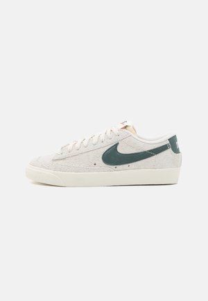 Bež nubuk tenisice s zelenim Nike swooshom, bijelim vezicama, teksturiranim gornjim dijelom i kremastom gumenom potplatom. Minimalistički dizajn, niska silueta.
