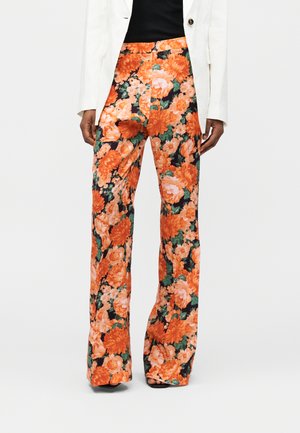 Oseba nosi široke hlače z oranžno in roza cvetličnim vzorcem, združene s črnim topom in belim blazerjem.